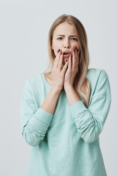 Shocked stunned blonde european woman with opened mouth, holding hands on cheeks, shocked by news she heard. Frightened terrified beautiful female model. Negative emotions
